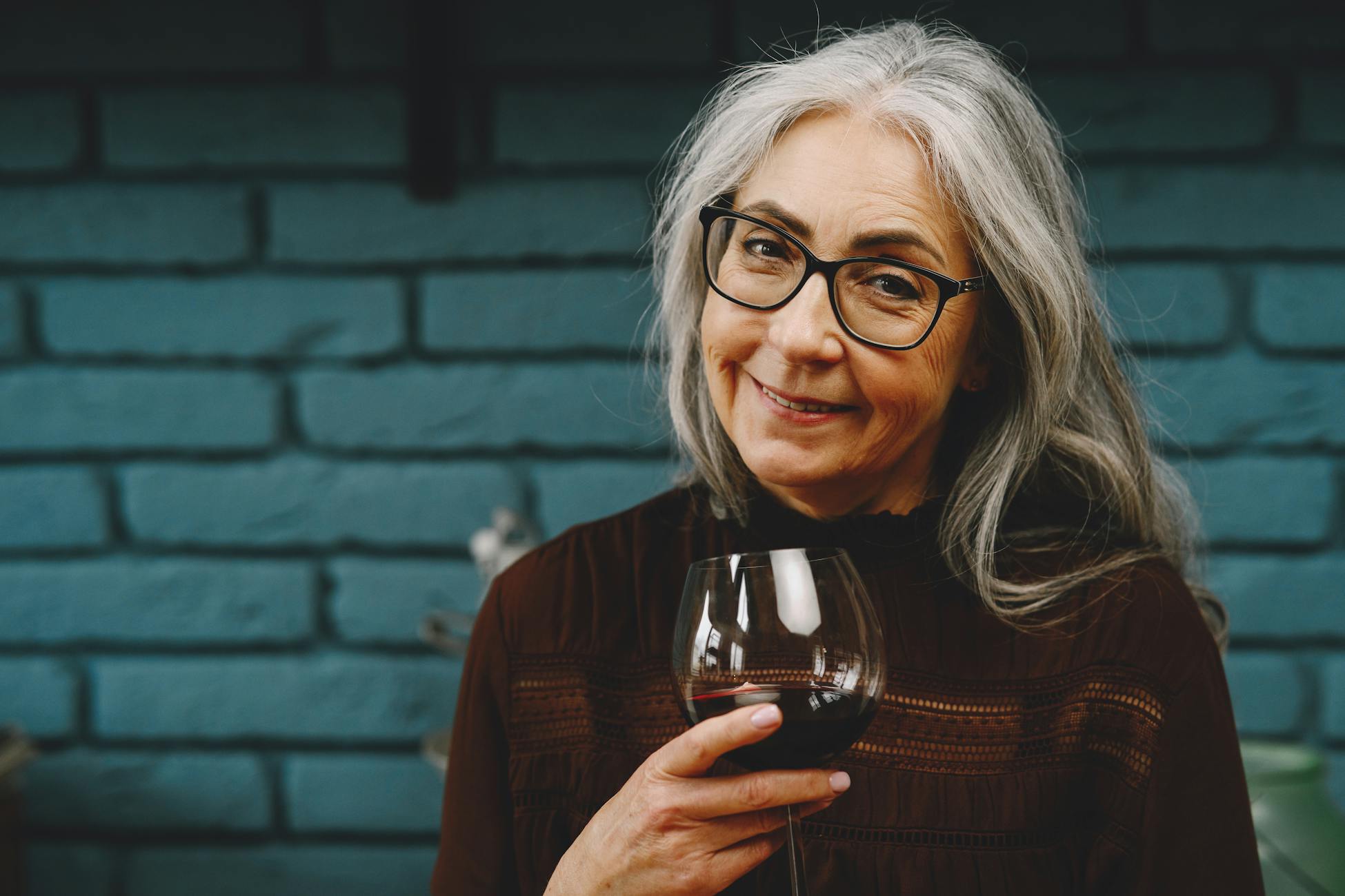 Smiling senior woman with eyeglasses holding a wine glass indoors against a brick wall.