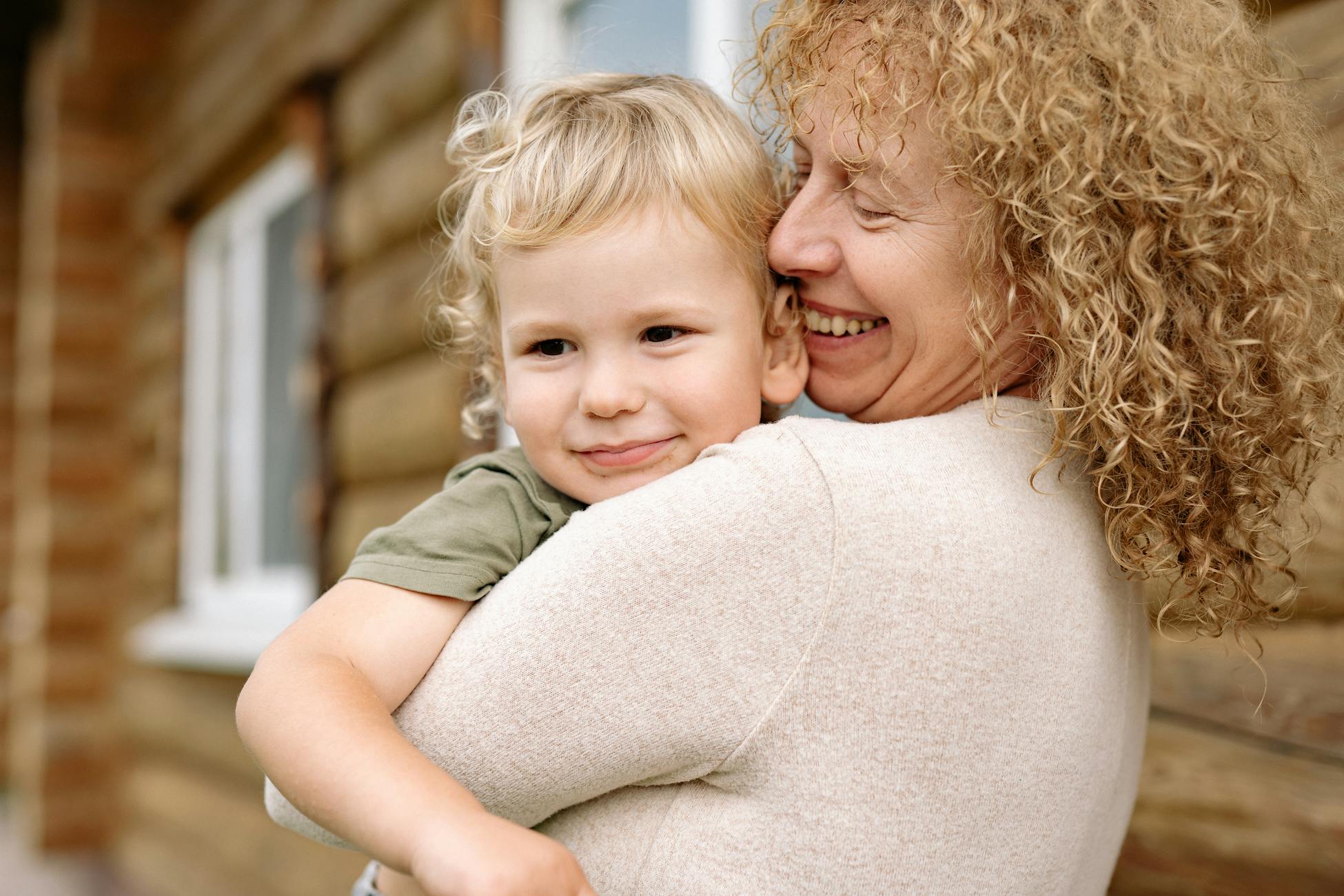 A cheerful senior woman embraces her smiling grandchild outside a wooden house, showcasing family love.