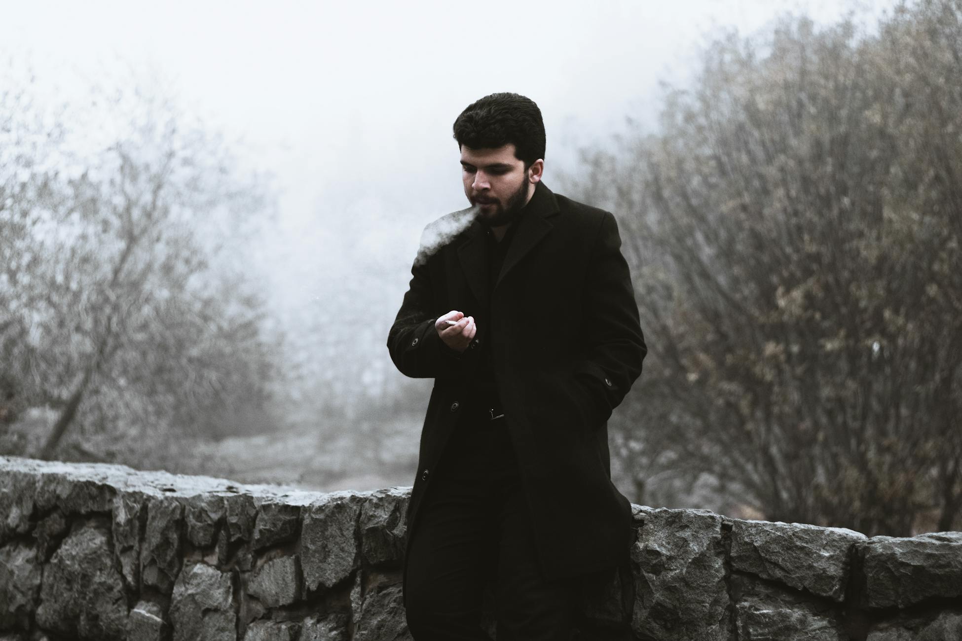 Portrait of a man smoking by a stone wall in a foggy outdoor setting.