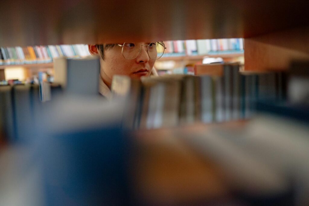 Young man wearing glasses studying intently between library shelves filled with books.