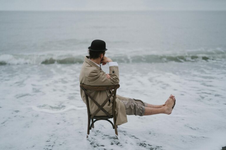Man in coat and hat sitting on a chair at the edge of the ocean, lost in thought.