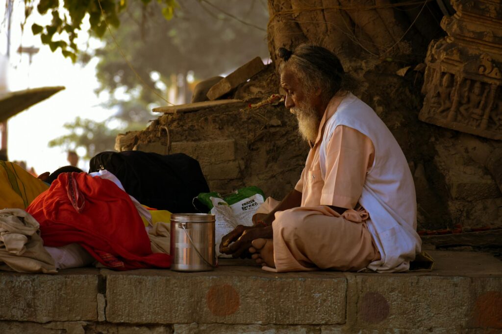 Elderly man sits peacefully in meditation beside a tree outdoors, surrounded by colorful fabric.