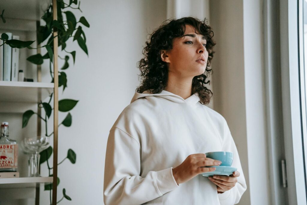 Young woman in hoodie holds cup by window, looking thoughtful.