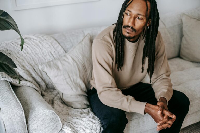 A man with dreadlocks sitting thoughtfully on a couch indoors, exuding a calm and pensive mood.