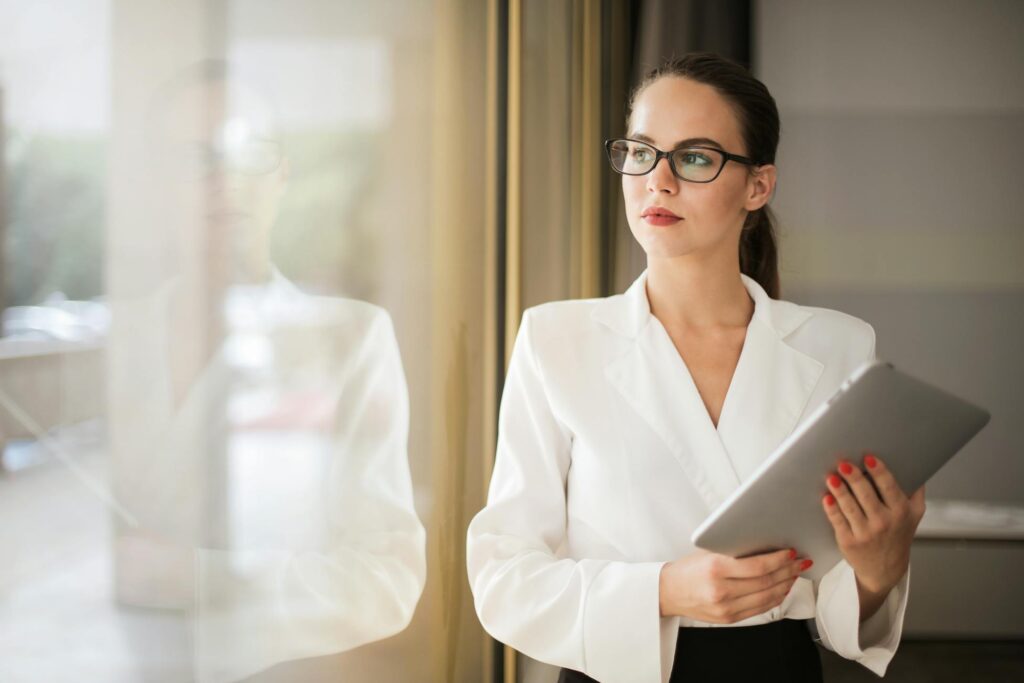 Businesswoman in formal attire thinking beside a window with a tablet.