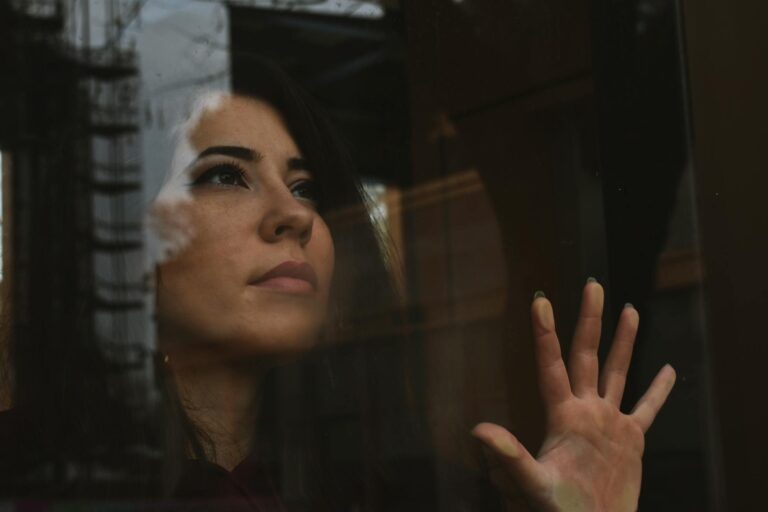 A woman reflects thoughtfully while looking out a window with her hand touching the glass.
