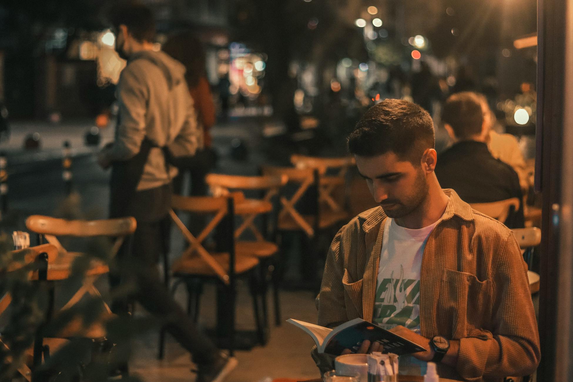 A man engrossed in a book while sitting at an outdoor cafe during the night.