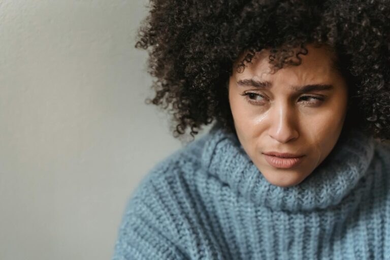 Crying ethnic female with curly hair looking away while thinking over problem and crying near white wall in studio