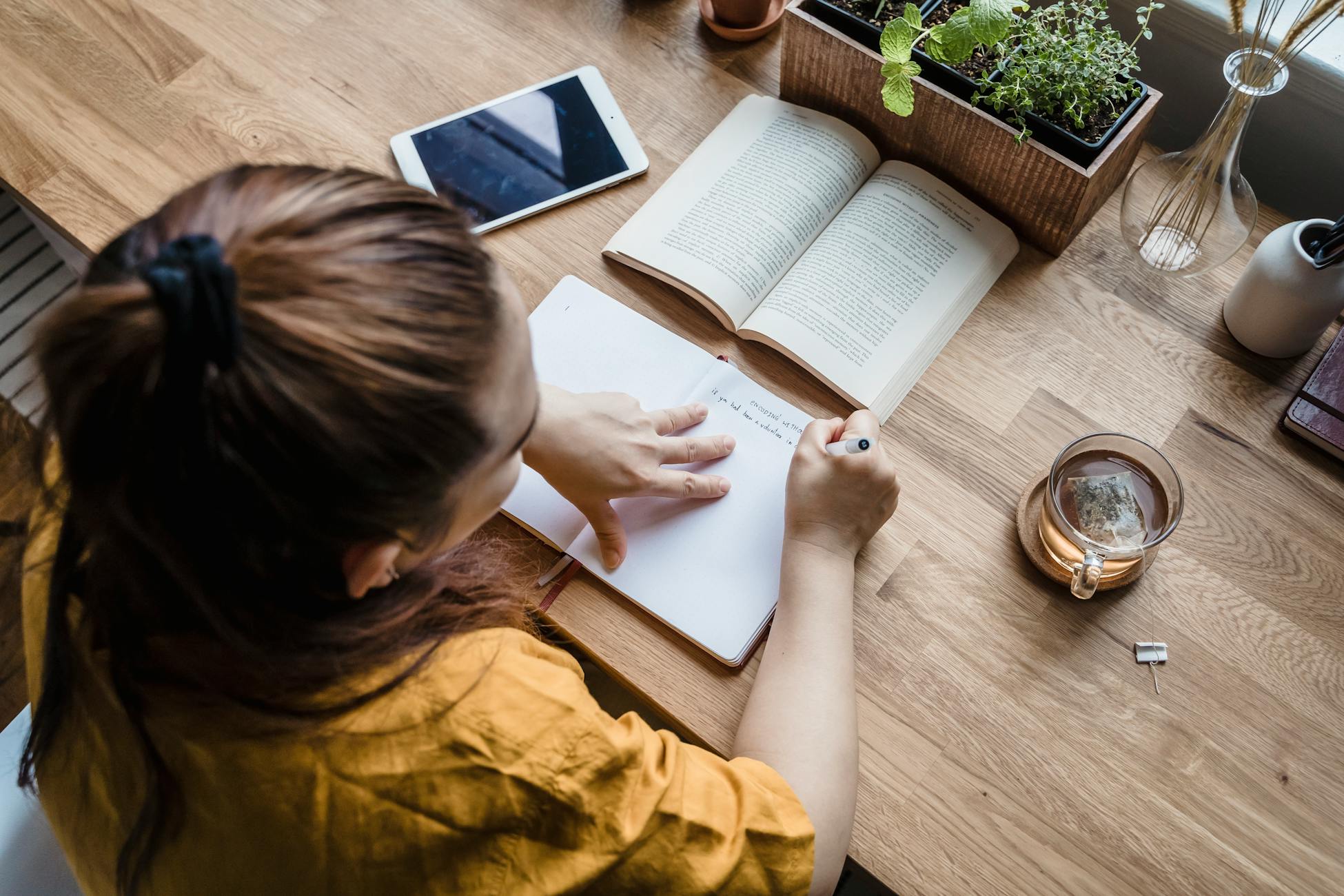 A woman writing in a notebook at a wooden desk with books, tea, and tablet.