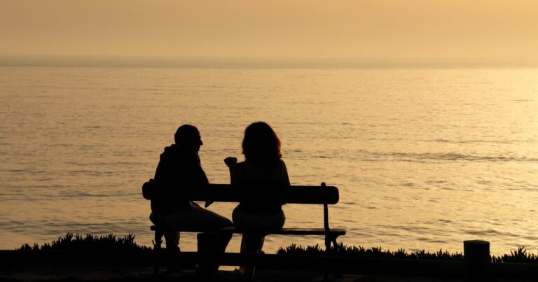 Two people sit talking on a bench by the sea at sunset, in silhouette