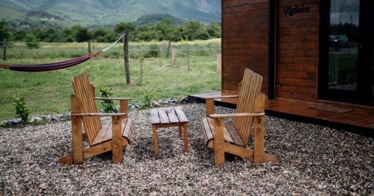 Two wooden Adirondack chairs facing a small table on gravel, with green hills behind
