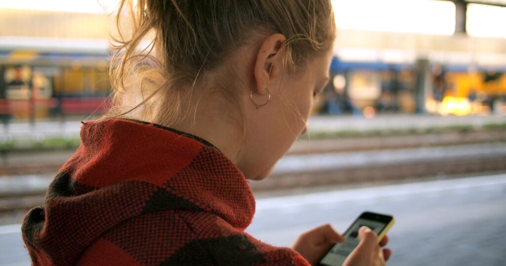 A young woman in a red plaid coat, seen from behind, typing on her phone while waiting on a train platform in late afternoon light.