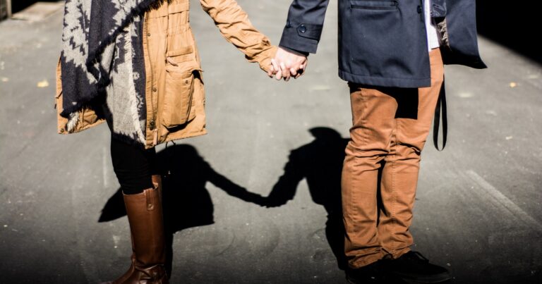 Two people in warm coats holding hands on a sunlit street, their joined shadow stretching across the pavement