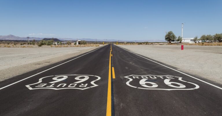 A freshly paved, empty desert highway running straight toward the horizon under a wide open sky, with large Route 66 shield markings painted on the asphalt