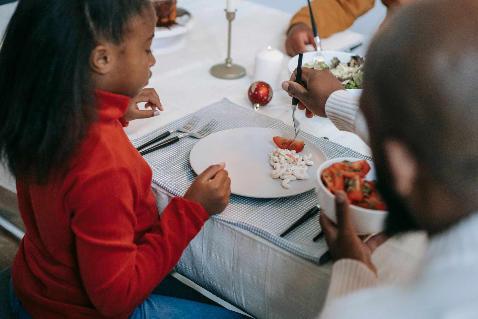 From above of crop anonymous ethnic parent serving pasta salad and tomato slice to girl while celebrating New Year at home
