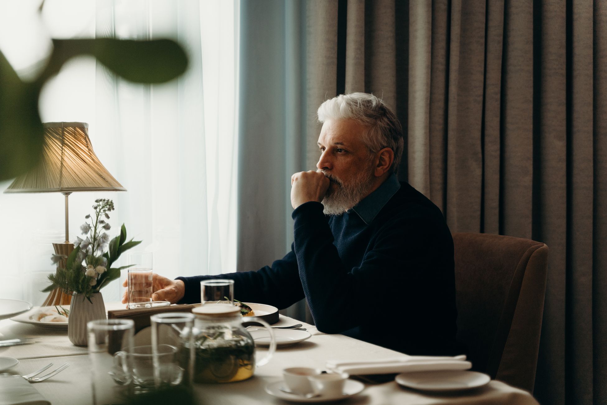 Thoughtful senior man with gray hair sitting at a dining room table, lost in thought.