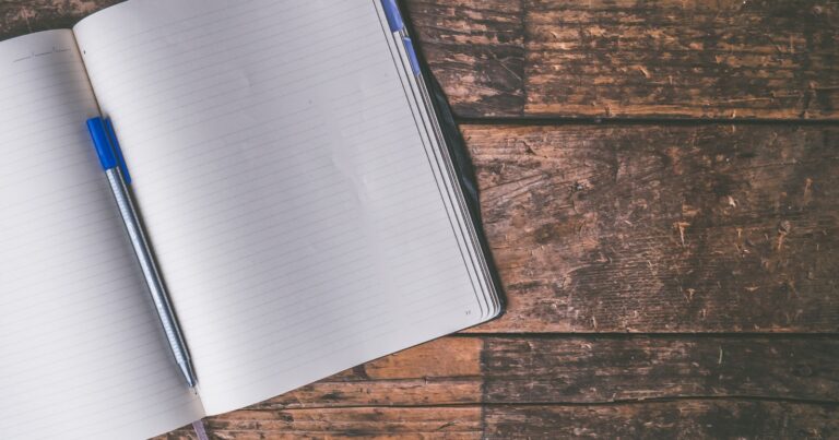 An open lined notebook with a ballpoint pen lying across it on a dark wooden table