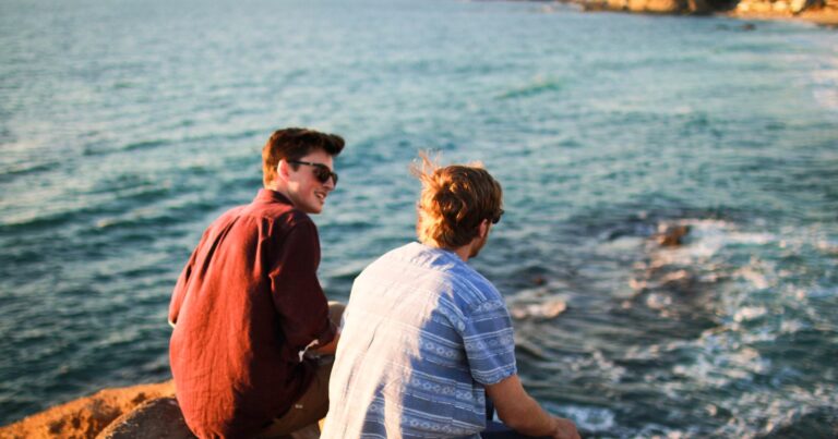 Two young men sit side by side on rocks by the sea in evening light, both facing the water.