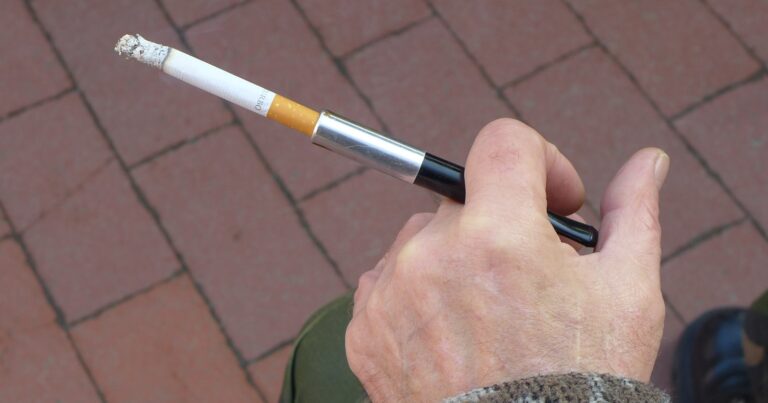 A person's hand resting on a knee, holding a lit cigarette in a slim holder, brick paving below