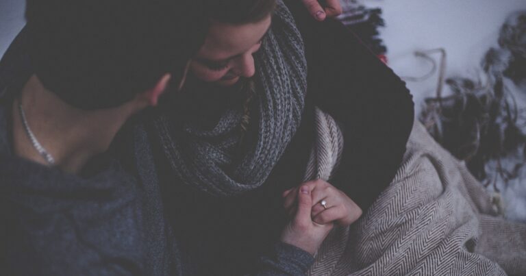 Two people sitting close together under knitted blankets, hands linked, faces mostly turned away