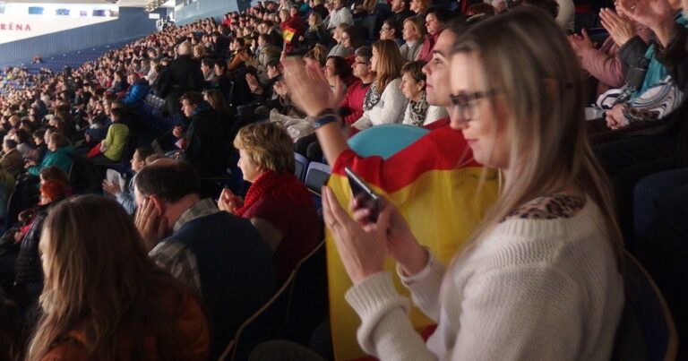 Spectators in the stands at a figure skating championship, a woman in the foreground in profile holding a flag and watching the ice