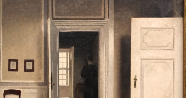 A woman in a dark dress stands with her back turned in the doorway of a pale, quiet interior, beside a bright window