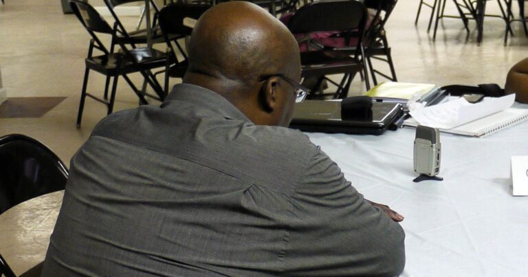 A man seen from behind sits at a round table across from an interviewer, with a small voice recorder and an open notebook between them and a hand-lettered sign reading Oral History Table.