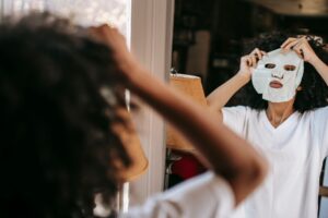 Concentrated African American female in domestic cloth putting moisturizing sheet mask while standing near mirror at home in daylight