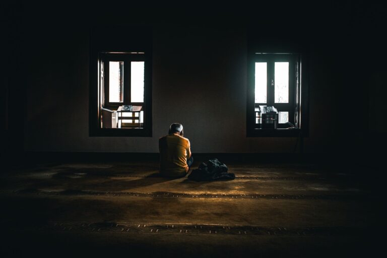 A solitary figure sits in contemplative silence in a dimly lit room with soft natural light.